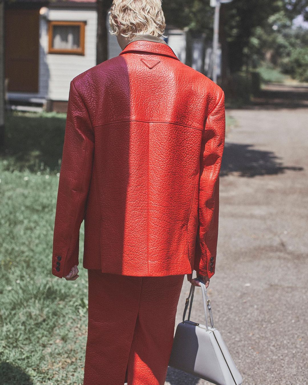 Back view of an adult wearing a red textured suit and carrying a grey bag in an outdoor environment with grass and buildings.