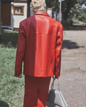 Back view of an adult wearing a red textured suit and carrying a grey bag in an outdoor environment with grass and buildings.