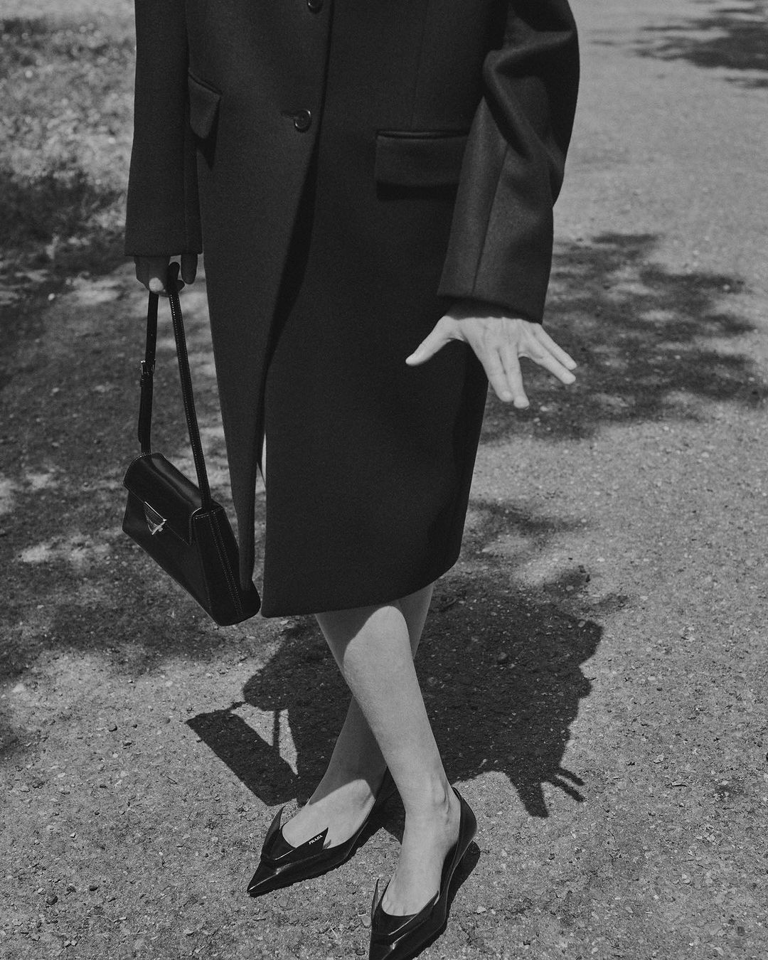 Black and white fashion shot features an adult female wearing a dark coat and shoes while holding a handbag on a sidewalk.
