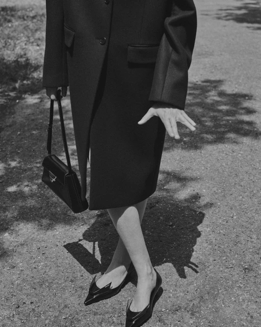 Black and white fashion shot features an adult female wearing a dark coat and shoes while holding a handbag on a sidewalk.