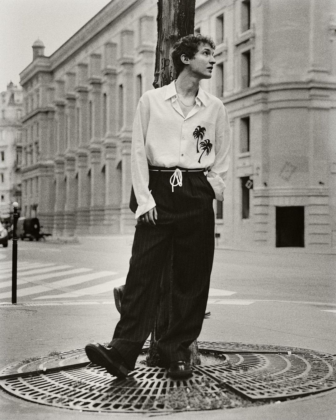 Monochrome portrait of a young adult man with curly hair leaning against a pole in a city street wearing a patterned button-down shirt.