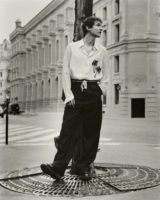 Monochrome portrait of a young adult man with curly hair leaning against a pole in a city street wearing a patterned button-down shirt.