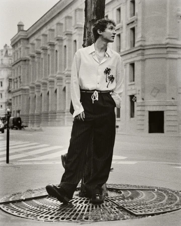 Monochrome portrait of a young adult man with curly hair leaning against a pole in a city street wearing a patterned button-down shirt.