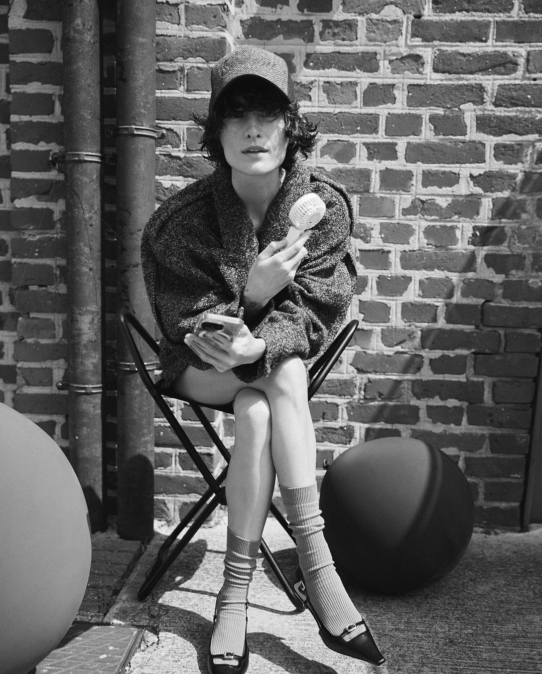 Black and white portrait of a woman sitting on a chair, holding a brush and phone, wearing an overcoat, socks, and flats in an urban setting.