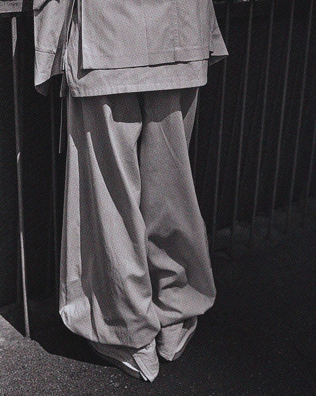 Monochrome fashion detail: lower half of a person in wide-legged pants and jacket, standing against a dark, barred background outdoors.