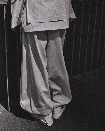 Monochrome fashion detail: lower half of a person in wide-legged pants and jacket, standing against a dark, barred background outdoors.