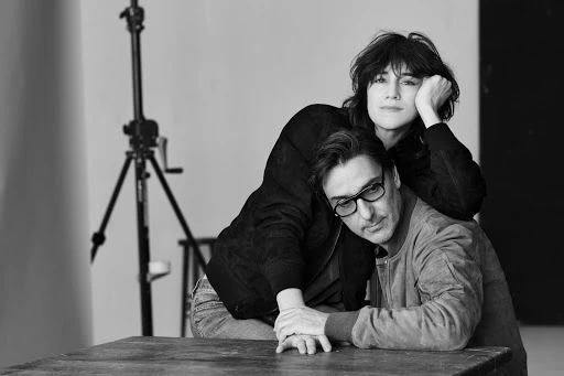 Black and white portrait of a woman resting her head on a man wearing glasses, seated at a dark wooden table in a studio.