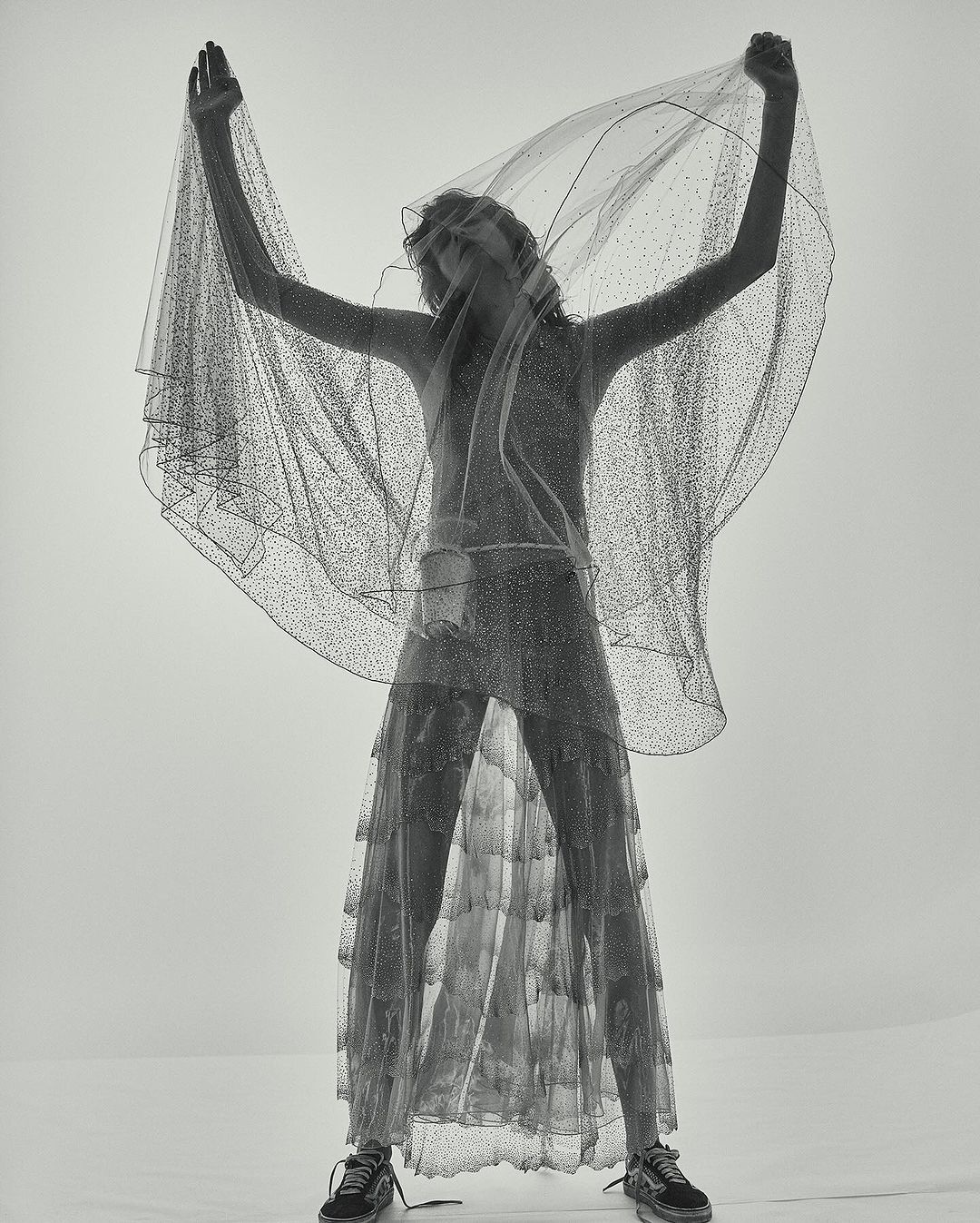 A slender woman in a semi-transparent glittery dress and sneakers poses under a sheer veil in a bright, minimalist studio setting.