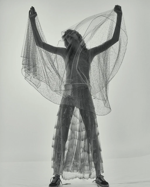 A slender woman in a semi-transparent glittery dress and sneakers poses under a sheer veil in a bright, minimalist studio setting.