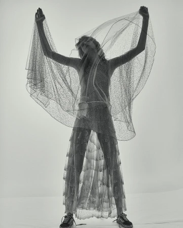 A slender woman in a semi-transparent glittery dress and sneakers poses under a sheer veil in a bright, minimalist studio setting.