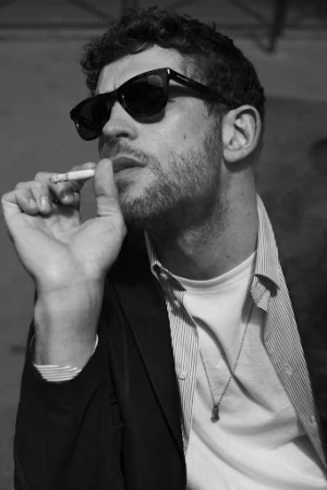 Black and white portrait of a man in sunglasses holding a cigarette, wearing a striped shirt and dark jacket by photographer Stefano Galuzzi.