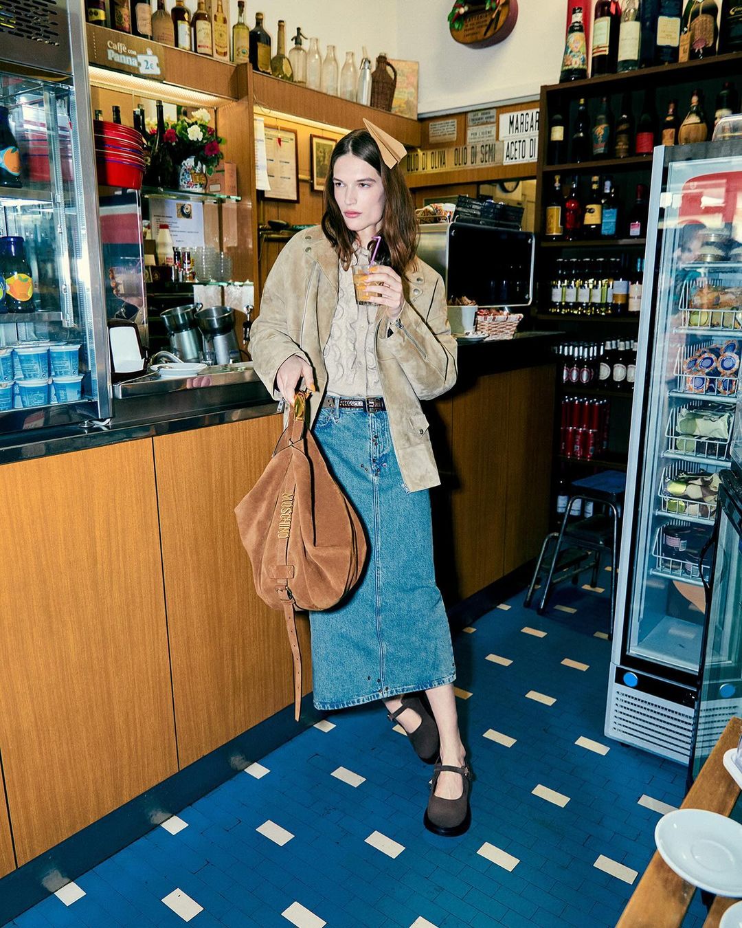 A woman in a beige jacket and denim skirt holds a drink and handbag in a vintage-style cafe setting, with blue patterned flooring.