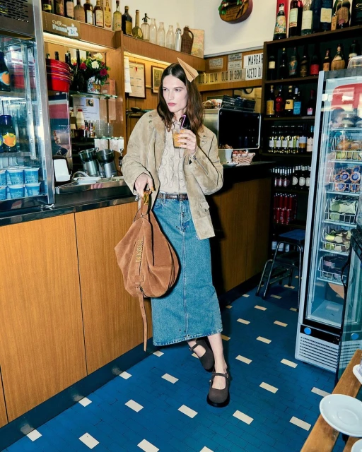 A woman in a beige jacket and denim skirt holds a drink and handbag in a vintage-style cafe setting, with blue patterned flooring.