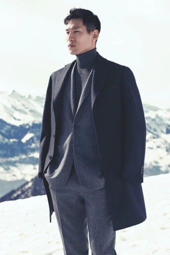 Asian man in gray suit and black coat standing in a snowy mountain landscape on a bright day; fashion advertising, apparel.