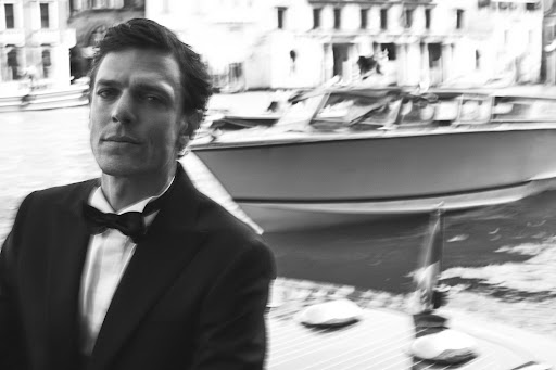 Monochrome portrait of a man in a tuxedo with a bow tie, on a boat, against blurred Venice buildings and water background.
