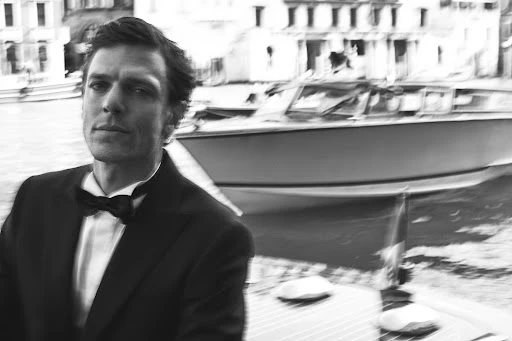Monochrome portrait of a man in a tuxedo with a bow tie, on a boat, against blurred Venice buildings and water background.