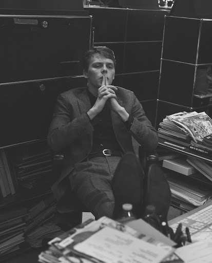 Man in suit sitting at a cluttered desk with hands together and feet on the desk in a black and white indoor shot with filing cabinets.