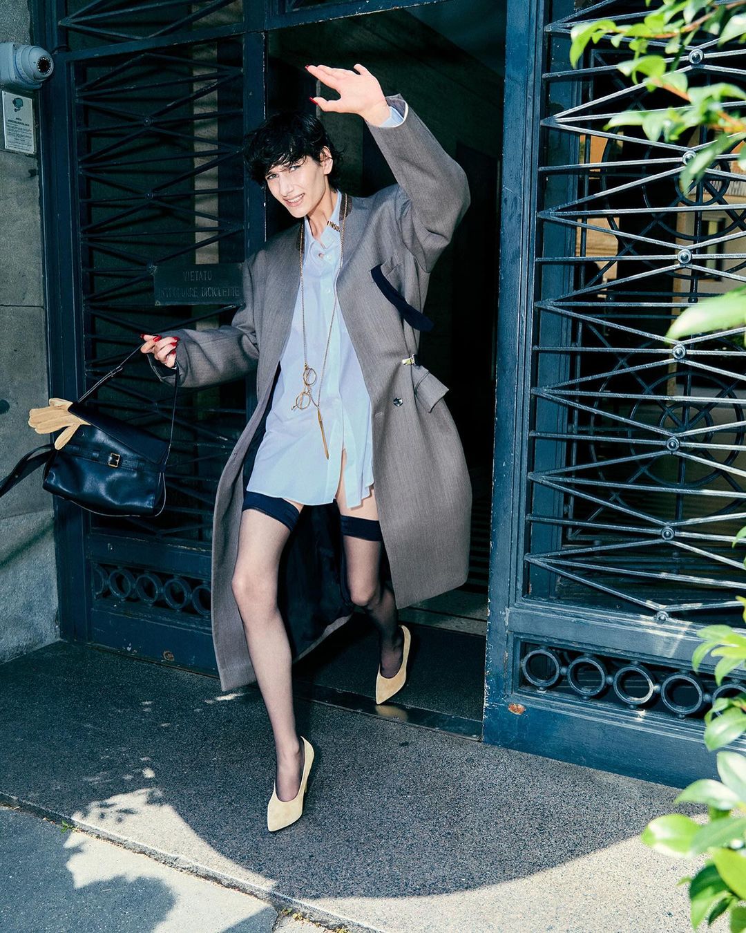 Person exiting a doorway, wearing an oversized grey coat, sheer stockings and light-colored heels, carrying a black bag in daytime.