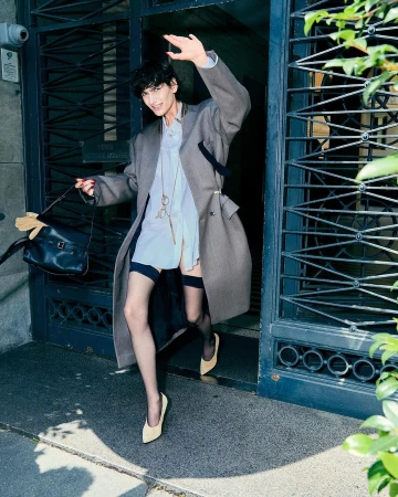 Person exiting a doorway, wearing an oversized grey coat, sheer stockings and light-colored heels, carrying a black bag in daytime.