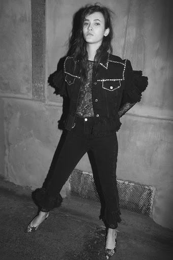 Black and white portrait of a woman in a black lace top, jacket, and trousers with fur trim posing against a textured grey wall.