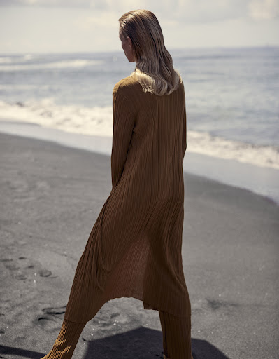 Model standing on a dark sand beach wearing a long brown ribbed knit cardigan and matching trousers