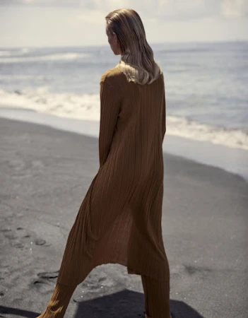 Model standing on a dark sand beach wearing a long brown ribbed knit cardigan and matching trousers
