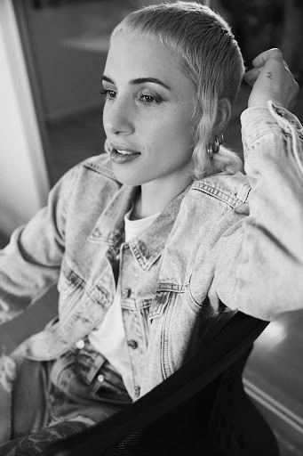 Portrait of a young adult female with a buzzcut hairstyle, wearing a denim jacket, in a black and white indoor shot.