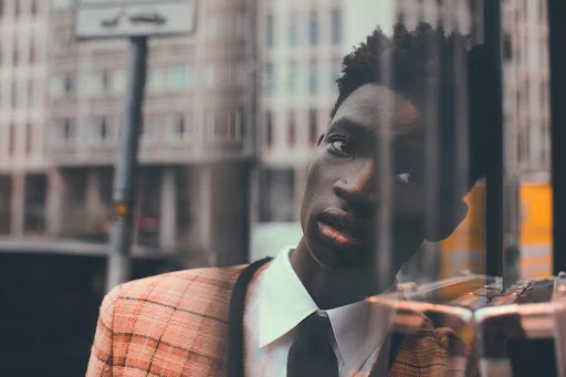 Close-up portrait of a Black man wearing a plaid jacket, white shirt, and black tie, reflected in a glass window.