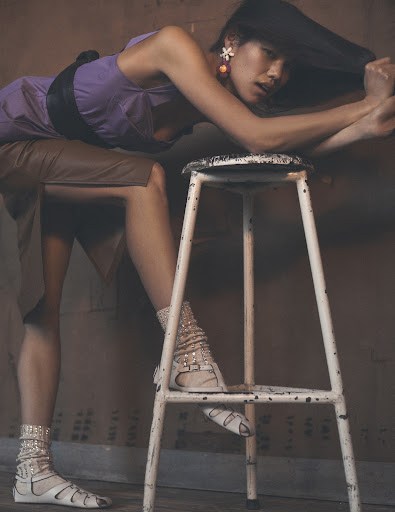 An adult woman in a purple dress, gladiator sandals, and white ankle socks rests her arms on a white metal stool inside a studio.