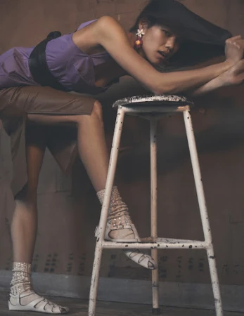 An adult woman in a purple dress, gladiator sandals, and white ankle socks rests her arms on a white metal stool inside a studio.