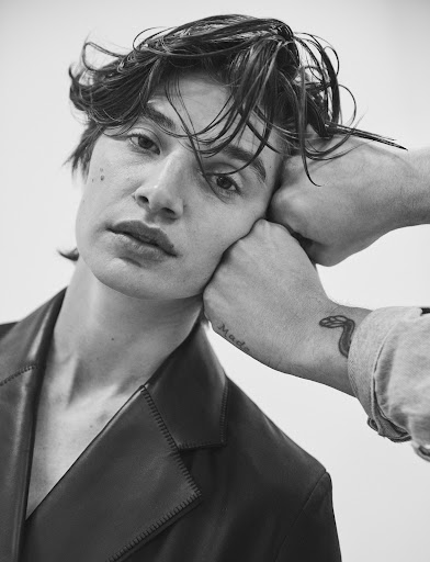 Black and white portrait of a young adult man with dark, wet hair and fair skin, with an arm resting near his face in a studio setting.