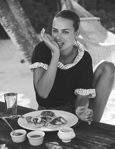 Black and white portrait of a woman sitting at a table with food, wearing a ruffled top, with a hammock in the background.