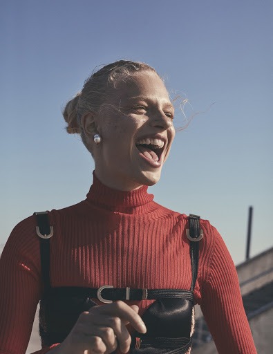 A woman smiles in a red turtleneck and black harness against a blue sky, captured in a medium shot showcasing a fashion-forward style.