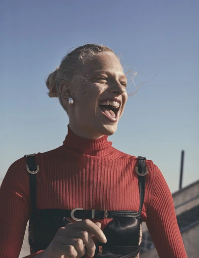 A woman smiles in a red turtleneck and black harness against a blue sky, captured in a medium shot showcasing a fashion-forward style.