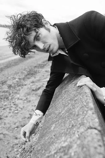 Black and white fashion shot of a young adult male model with curly hair, wearing a coat, posing near a concrete barrier outdoors.