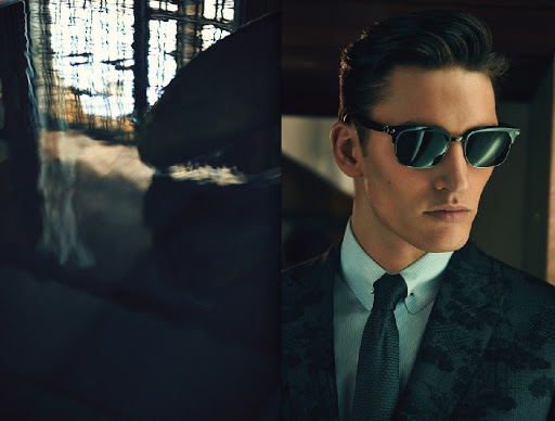 A sharply dressed man in a suit and sunglasses poses indoors, bathed in ambient light, showcasing classic style and sophistication.