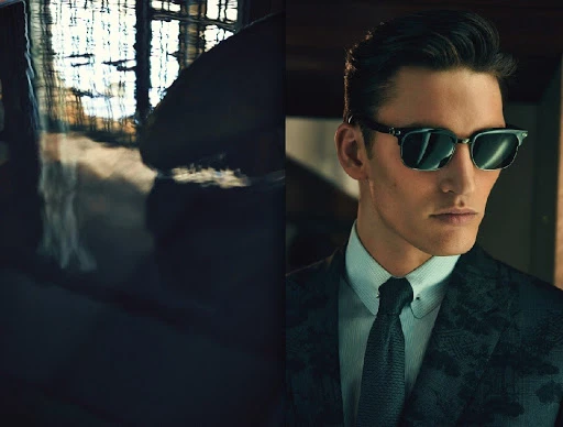 A sharply dressed man in a suit and sunglasses poses indoors, bathed in ambient light, showcasing classic style and sophistication.