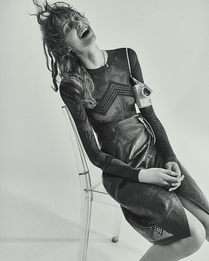 Monochromatic studio shot of a laughing woman in a patterned top and skirt sitting on a transparent chair with an old-fashioned camera.