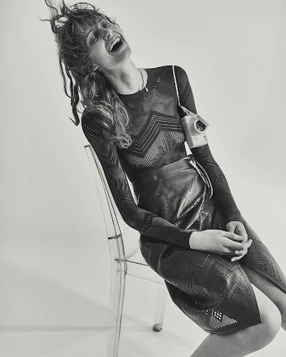 Monochromatic studio shot of a laughing woman in a patterned top and skirt sitting on a transparent chair with an old-fashioned camera.