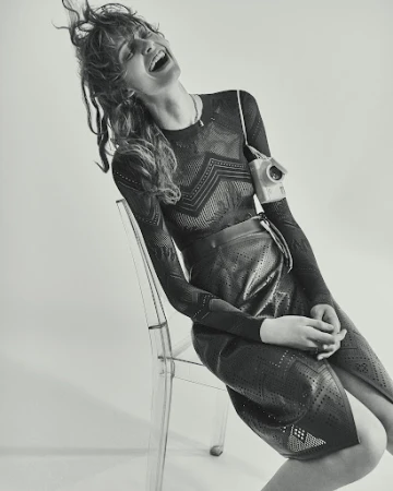Monochromatic studio shot of a laughing woman in a patterned top and skirt sitting on a transparent chair with an old-fashioned camera.