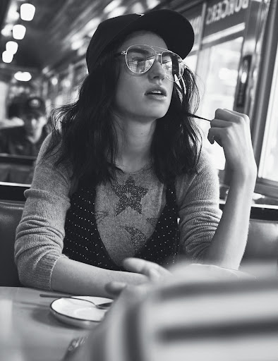 Black and white shot of a stylish young woman wearing glasses and a cap, seated at a booth in a diner.