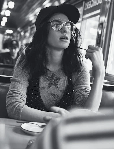 Black and white shot of a stylish young woman wearing glasses and a cap, seated at a booth in a diner.