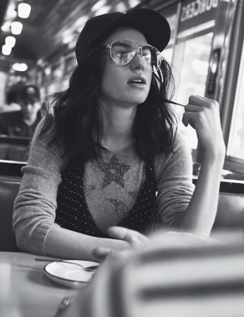 Black and white shot of a stylish young woman wearing glasses and a cap, seated at a booth in a diner.