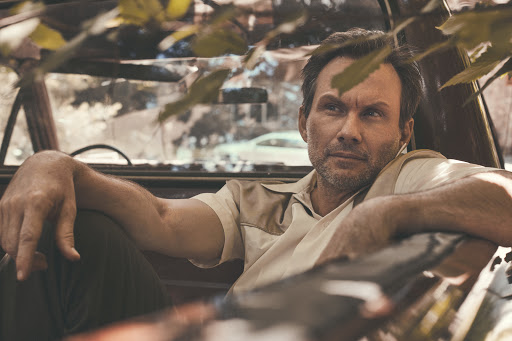 Adult male relaxes in old car, wearing a vintage-style shirt; sunlight filters through the leaves to add warmth to the scene.
