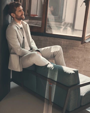 Man seated on a teal sofa wearing a light grey suit jacket and trousers near a window by Stefano Galuzzi