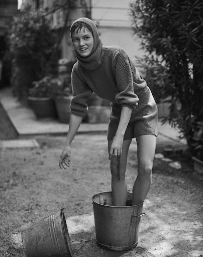 A woman stands in a bucket of liquid in a black and white, mid-shot, outside, wearing knitted sweater and shorts.