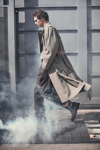 A man wearing a trench coat is walking through a cloud of smoke near a gray metal industrial gate, in an outdoor urban setting.