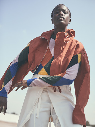 Woman in a colorful jacket poses against a light blue sky. The jacket has geometric patterns and is cropped above white wraparound pants.