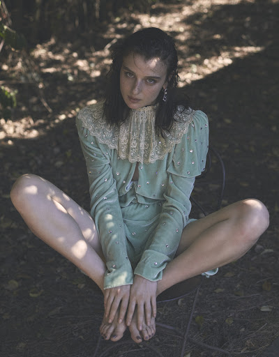 A light-skinned woman with dark hair is sitting barefoot outdoors in a chair wearing a light green embellished outfit.