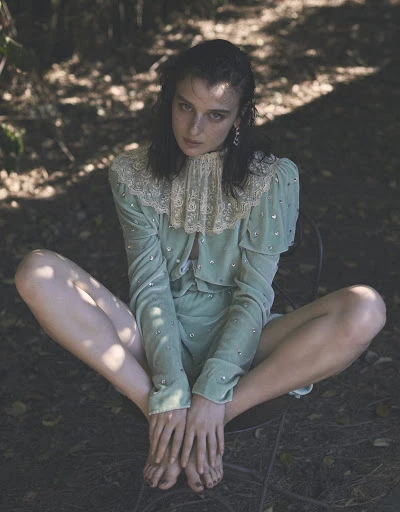 A light-skinned woman with dark hair is sitting barefoot outdoors in a chair wearing a light green embellished outfit.